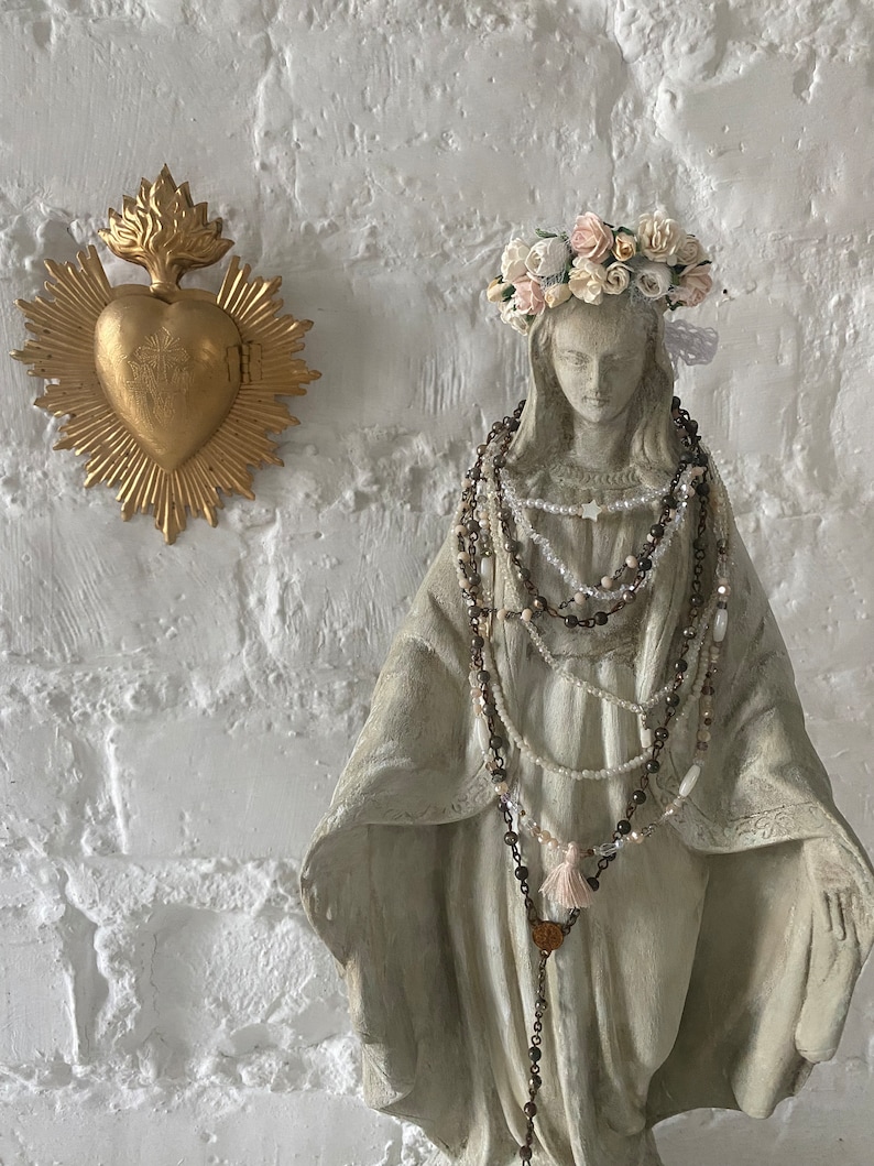 May include: A white statue of the Virgin Mary wearing a crown of white and pink flowers. She is adorned with multiple beaded necklaces, including a rosary with a tassel. The statue is set against a white brick wall with a gold heart-shaped religious icon hanging above.