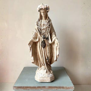 Puede incluir: Estatua beige de una mujer con corona floral y rosarios. La estatua est&aacute; sobre una superficie azul claro. Los rosarios incluyen un colgante en forma de coraz&oacute;n. Expresi&oacute;n serena.