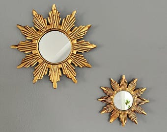 May include: Two gold sunburst mirrors of different sizes on a grey wall. Each mirror has a round, reflective surface surrounded by a sunburst design. The larger mirror is approximately 30 cm in diameter.