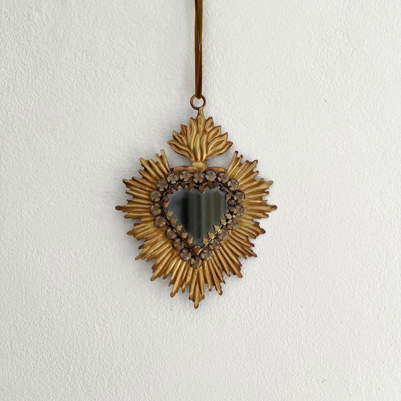 May include: A gold-colored, heart-shaped wall mirror with a sunburst design. The mirror is framed with small, clear stones and has a loop for hanging. The mirror is suspended from a thin, brown ribbon.