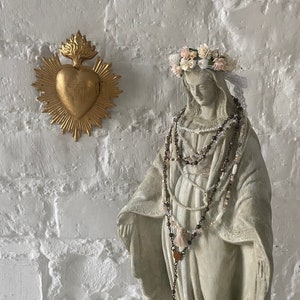 May include: A white plaster statue of the Virgin Mary wearing a crown of white and pink flowers and multiple beaded necklaces. The statue is standing in front of a white brick wall with a gold heart-shaped religious icon.