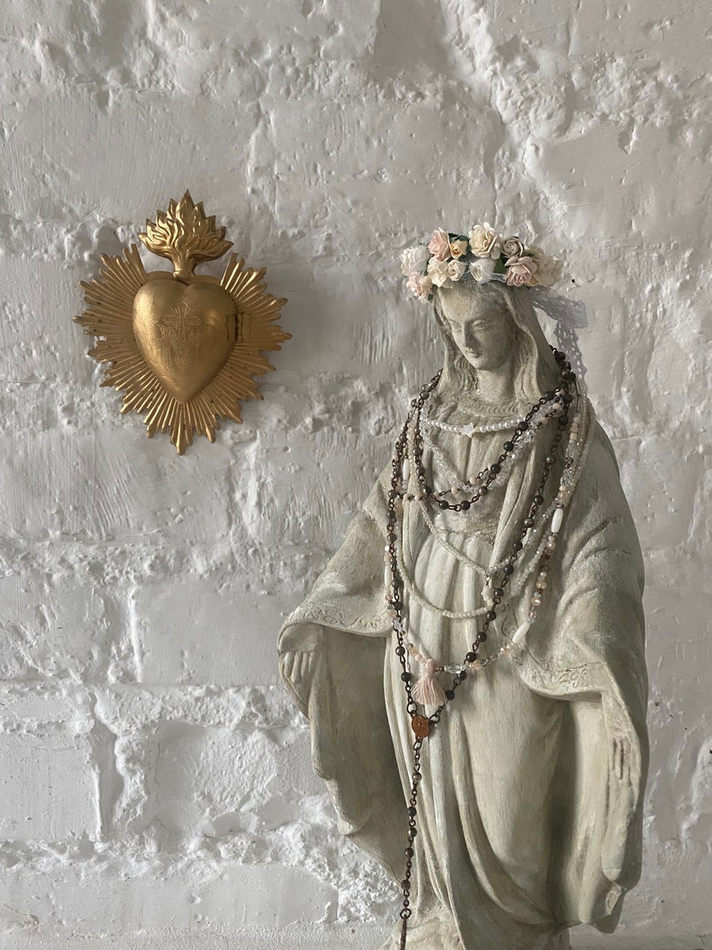 May include: A white plaster statue of the Virgin Mary wearing a crown of white and pink flowers and multiple beaded necklaces. The statue is standing in front of a white brick wall with a gold heart-shaped religious icon.