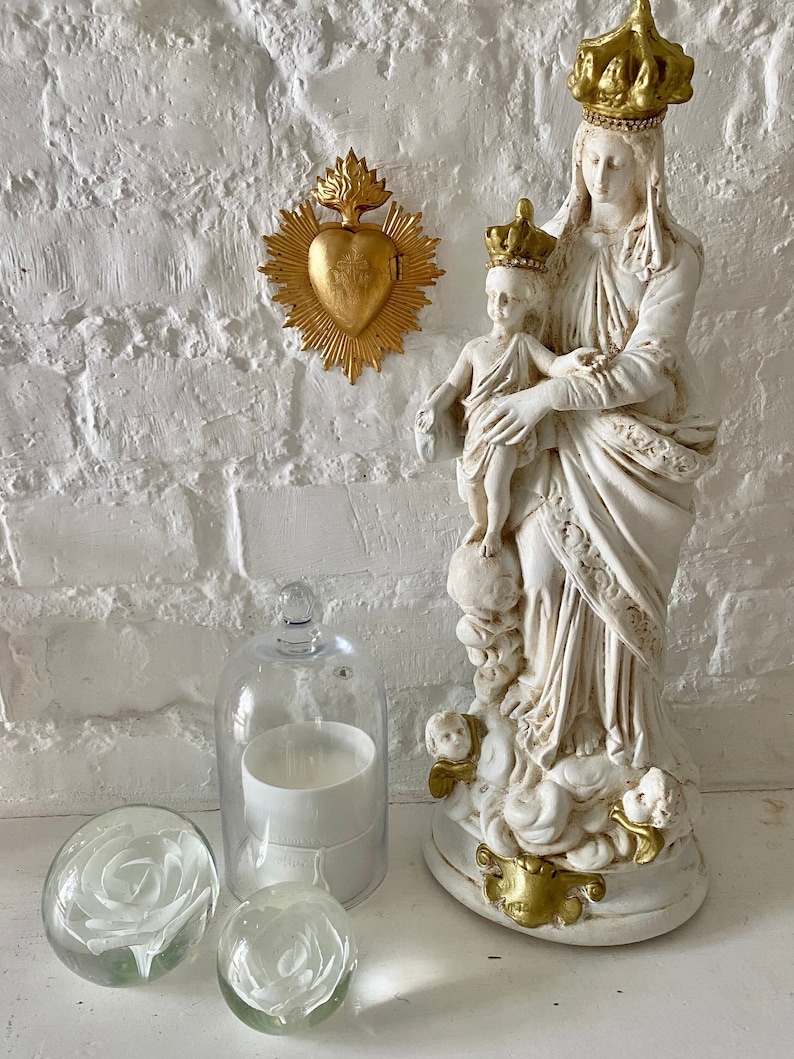May include: White ceramic statue of the Virgin Mary holding a baby Jesus. The statue is adorned with gold accents and a crown. The statue is displayed on a white shelf with a gold heart ornament and glass orbs.