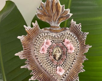 May include: Heart-shaped religious wall decoration featuring an image of the Virgin Mary and child. The heart is decorated with rhinestones and pink flowers. The frame has a gold and white distressed finish. The item is approximately 15 cm in height.