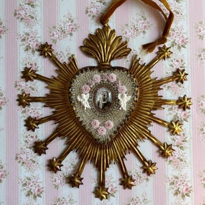 May include: A gold-colored, heart-shaped religious wall hanging with a central image, surrounded by radiating rays and starbursts. The heart is embellished with small pink roses, beads, and two white angel figures. A brown ribbon is attached for hanging.
