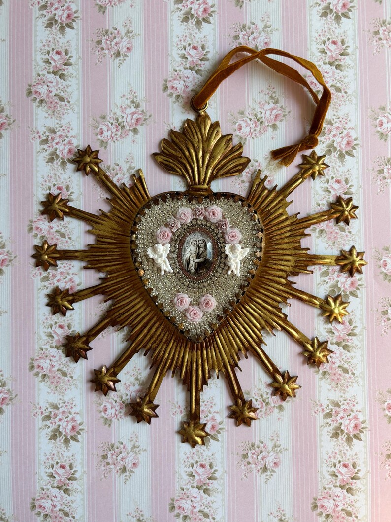 May include: A gold-colored, heart-shaped religious wall hanging with a central image, surrounded by radiating rays and starbursts. The heart is embellished with small pink roses, beads, and two white angel figures. A brown ribbon is attached for hanging.