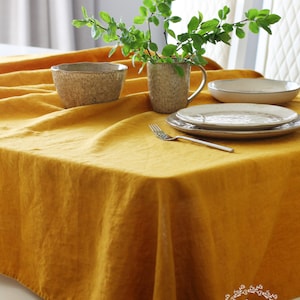 Può includere: Una tovaglia di lino gialla con una ciotola, una tazza e piatti sopra. La tovaglia è su un tavolo bianco con due sedie bianche sullo sfondo. Il testo "Linen Home Studio" è visibile nell'angolo in basso a destra dell'immagine.