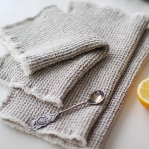 May include: Three waffle weave linen kitchen towels in a light gray color folded on a white surface with a silver spoon and two lemon halves.