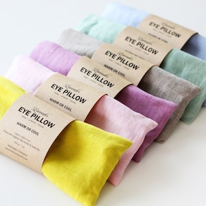 Pode incluir: Um conjunto de seis almofadas para os olhos em várias cores, incluindo amarelo, rosa, azul claro, verde e cinza. Cada almofada tem um rótulo castanho que diz "Lavender's Eye Pillow Warm or Cool".