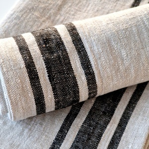 Può includere: Due asciugamani da tè in lino con strisce nere. Gli asciugamani sono piegati e impilati uno sopra l'altro.