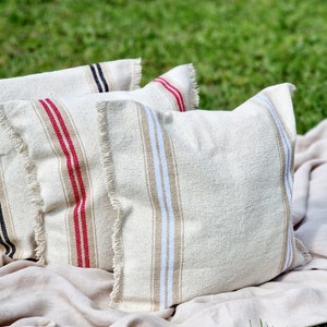 Può includere: Tre cuscini da lancio a righe con bordi sfrangiati. I cuscini sono beige con strisce rosse e bianche, beige con strisce blu e bianche e neri con strisce bianche. I cuscini sono disposti su una coperta beige.