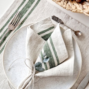 Peut inclure: Mise en place rustique avec une assiette blanche, une serviette en lin à rayures vertes et une branche de verdure. Une fourchette et une cuillère en argent sont sur la table, ainsi qu'un panier en osier rempli de noix. La serviette est nouée avec de la ficelle.