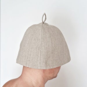 May include: A light beige sauna hat made of linen. The hat has a rounded crown and a small loop at the top. The hat is designed to protect the head from heat in a sauna.