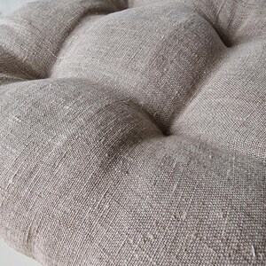 Extreme close-up of a natural oatmeal linen chair cushion, highlighting the rich, rustic woven texture of the fabric. The image shows the detailed slubs of the unbleached linen and the plump, tufted design of the seat pad.