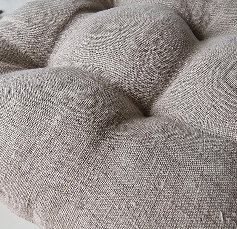Extreme close-up of a natural oatmeal linen chair cushion, highlighting the rich, rustic woven texture of the fabric. The image shows the detailed slubs of the unbleached linen and the plump, tufted design of the seat pad.