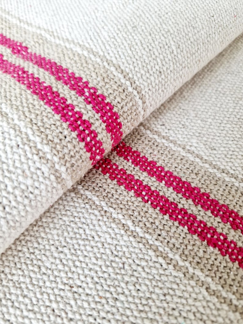 Pu&ograve; includere: Primo piano di un tessuto piegato di colore beige naturale. Due vivaci strisce fucsia attraversano il tessuto. La trama del materiale &egrave; visibile, creando un'estetica rustica e artigianale. Il tessuto sembra essere lino o una fibra naturale simile.
