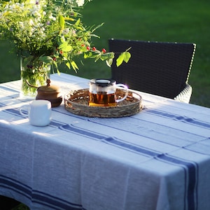 Horizontal view of a navy blue striped linen tablecloth on an outdoor garden table. The scene features a glass vase of wildflowers, a woven wicker tray with a glass teapot, and a white mug in the golden hour sunlight. Farmhouse style patio decor with a rustic, textured linen finish.