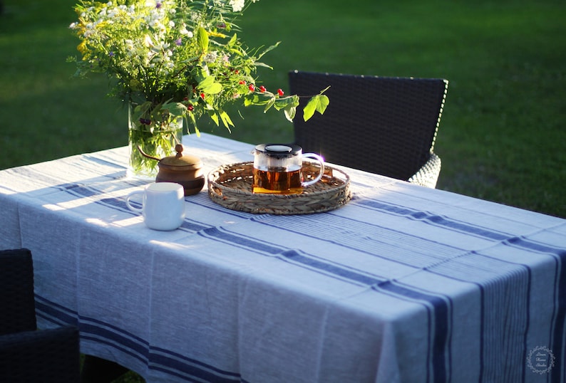 Horizontal view of a navy blue striped linen tablecloth on an outdoor garden table. The scene features a glass vase of wildflowers, a woven wicker tray with a glass teapot, and a white mug in the golden hour sunlight. Farmhouse style patio decor with a rustic, textured linen finish.
