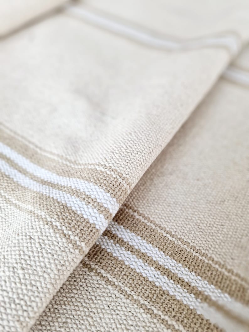 Pu&ograve; includere: Primo piano di tessuto di lino beige piegato con strisce bianche e beige. Il tessuto ha una trama strutturata e le strisce sono parallele tra loro. Le pieghe creano ombre e profondit&agrave;, evidenziando i toni naturali del materiale.