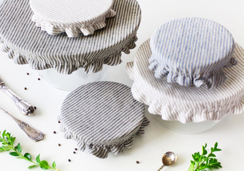 Set of reusable striped linen bowl covers with elastic edges. Eco-friendly zero waste fabric dish covers in grey and blue stripes, styled on a white table with vintage silver spoons and green sprigs. Sustainable kitchen gift and plastic wrap alternative.
