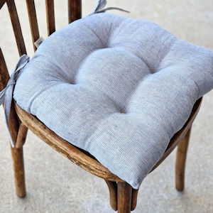 A square, tufted linen chair cushion with a light blue and white striped pattern, placed on a vintage wooden dining chair. The farmhouse style kitchen seat pad features integrated fabric ties secured to the back of the chair.
