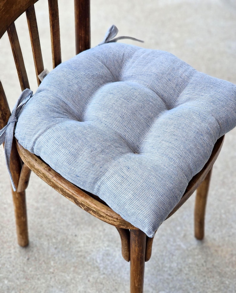 A square, tufted linen chair cushion with a light blue and white striped pattern, placed on a vintage wooden dining chair. The farmhouse style kitchen seat pad features integrated fabric ties secured to the back of the chair.