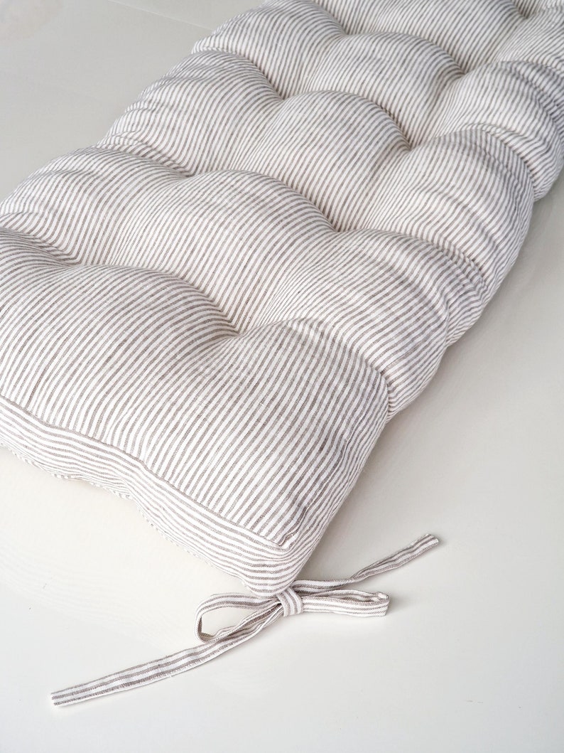 May include: A rectangular cushion with a striped pattern in shades of beige and white. The cushion is tufted, with a series of evenly spaced, rounded sections. A fabric tie is attached at one end. The cushion is designed for seating.