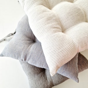 May include: A stack of four tufted seat cushions in various colors. The cushions are striped in shades of gray, blue, and off-white. The cushions are made of a soft fabric and have a rounded shape.