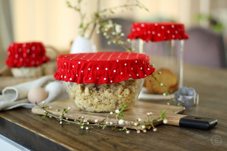 Vibrant red linen bowl covers with white polka dots. Set of reusable fabric jar lids with elastic edges on glass bowls, styled in a rustic kitchen with spring blossoms and eggs. Festive eco-friendly kitchen decor and zero-waste gift idea.