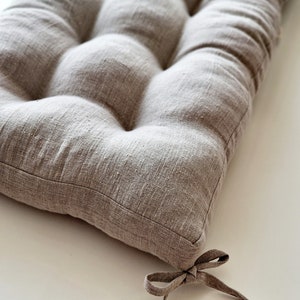 May include: A rectangular, tufted cushion in a neutral beige color. The cushion is made of a textured fabric and features a series of rounded, button-like tufts. A fabric tie is attached to one corner. The cushion is designed for seating.