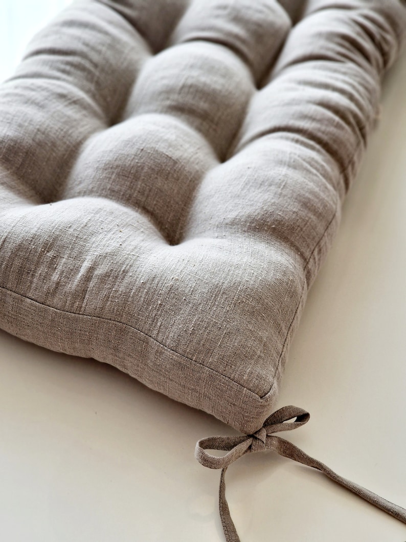 May include: A rectangular, tufted cushion in a neutral beige color. The cushion is made of a textured fabric and features a series of rounded, button-like tufts. A fabric tie is attached to one corner. The cushion is designed for seating.
