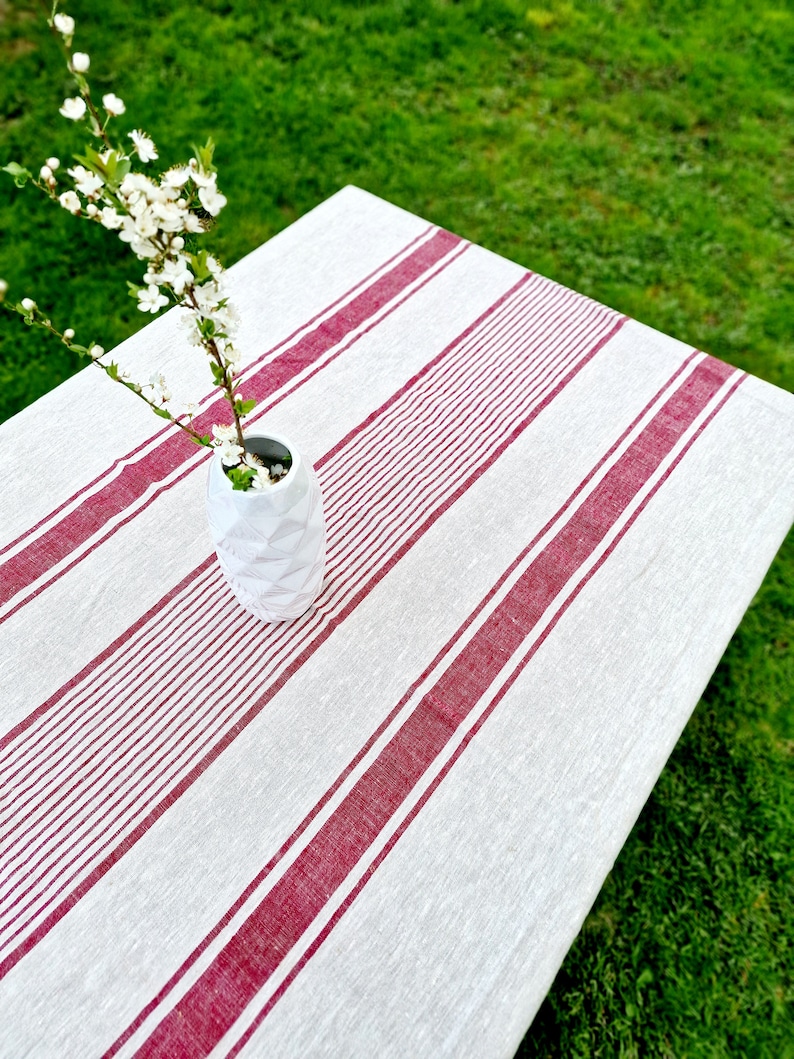 High-angle view of a rustic linen tablecloth with bold red grain sack stripes. A white textured vase with spring blossom branches sits on a garden table over green grass. The 100% natural flax fabric features a vintage French farmhouse aesthetic, perfect for outdoor dining or Easter table decor.