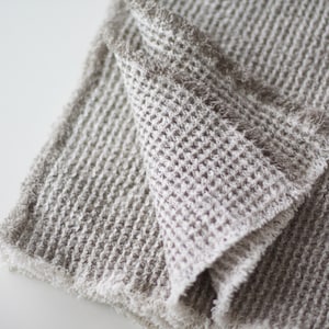 May include: A close-up of a soft, gray waffle weave throw blanket with a frayed edge. The blanket is folded in half and the texture of the fabric is visible.