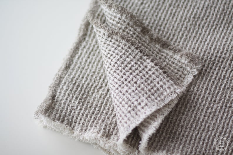 May include: A close-up of a soft, gray waffle weave throw blanket with a frayed edge. The blanket is folded in half and the texture of the fabric is visible.