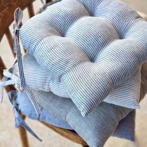 Stack of blue and grey striped linen chair pads with ties, resting on a vintage wooden chair. Handcrafted textured seat cushions for farmhouse style home decor and rustic kitchen seating.