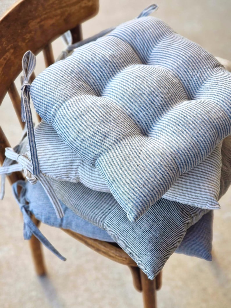Stack of blue and grey striped linen chair pads with ties, resting on a vintage wooden chair. Handcrafted textured seat cushions for farmhouse style home decor and rustic kitchen seating.