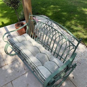 May include: A green metal garden bench with a striped cushion. The bench has a curved back and scrolled armrests. The cushion is light gray with dark green stripes. The bench is outdoors on a stone patio.