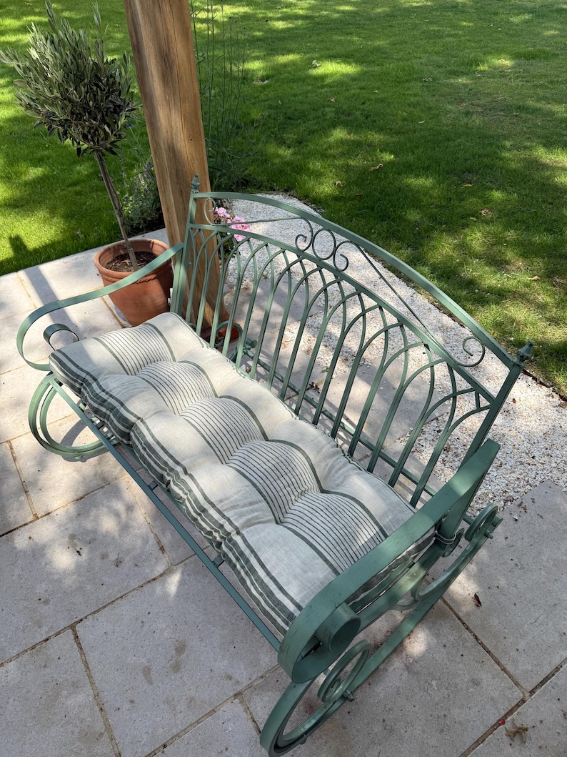 May include: A green metal garden bench with a striped cushion. The bench has a curved back and scrolled armrests. The cushion is light gray with dark green stripes. The bench is outdoors on a stone patio.