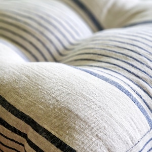 May include: Close-up of a tufted cushion with a cream-colored fabric and navy blue stripes. The stripes run horizontally across the cushion, creating a classic, nautical design. The cushion has a soft, textured appearance.