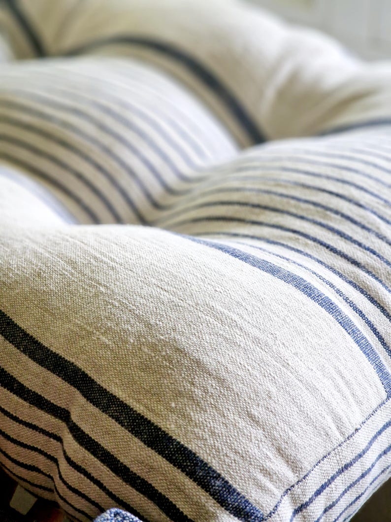 May include: Close-up of a tufted cushion with a cream-colored fabric and navy blue stripes. The stripes run horizontally across the cushion, creating a classic, nautical design. The cushion has a soft, textured appearance.