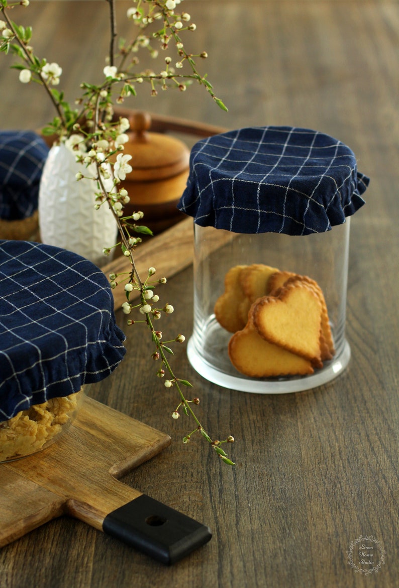 Dark navy blue checkered linen bowl and jar covers with a white grid pattern. Reusable fabric lids with elastic edges on glass containers with heart-shaped cookies. Cozy rustic kitchen decor and eco-friendly zero waste alternative to plastic wrap.
