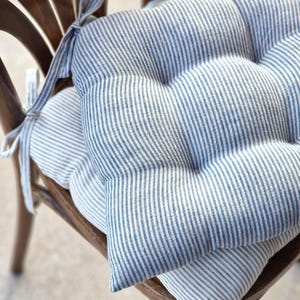 Two blue and grey striped linen chair cushions stacked on top of each other on a classic wooden dining chair. The top seat pad features vertical blue stripes on a natural light background, while the bottom cushion shows a matching stripe pattern. Both rustic kitchen pads are tufted and secured to the chair with integrated fabric ties.
