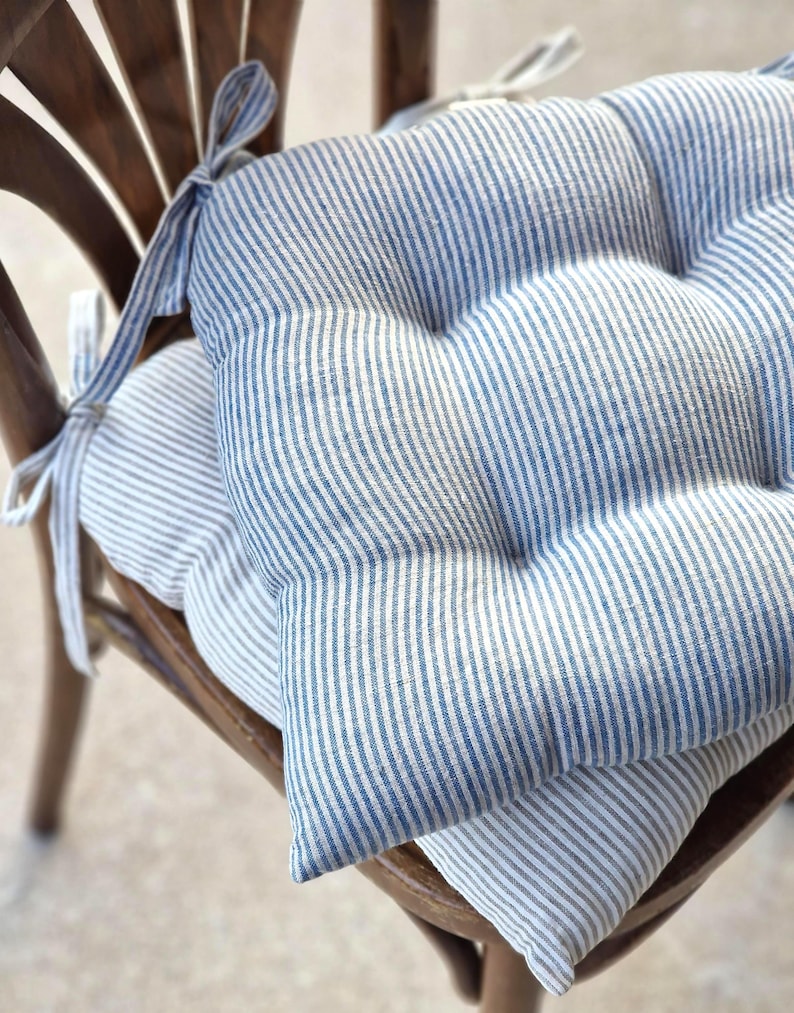 Two blue and grey striped linen chair cushions stacked on top of each other on a classic wooden dining chair. The top seat pad features vertical blue stripes on a natural light background, while the bottom cushion shows a matching stripe pattern. Both rustic kitchen pads are tufted and secured to the chair with integrated fabric ties.