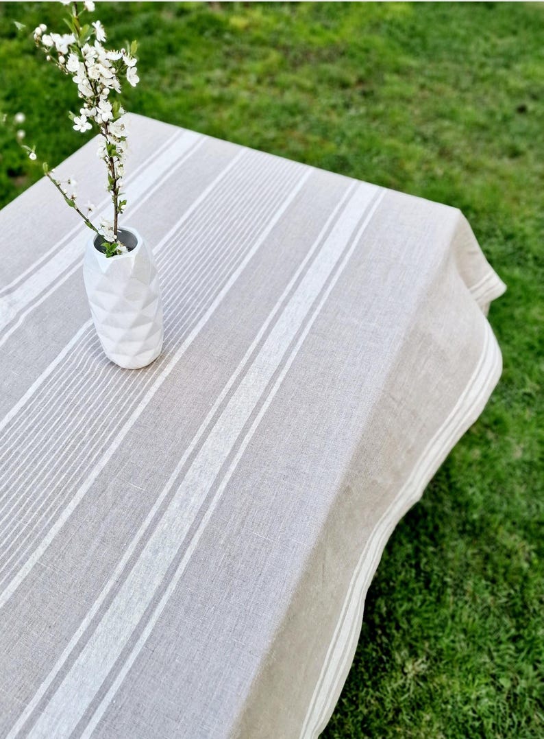High-angle view of a heavyweight oatmeal linen tablecloth with crisp white grain sack stripes. A white geometric vase with delicate spring blossoms sits on a garden table over green grass. This rustic, 100% natural flax tablecloth provides a vintage French farmhouse aesthetic for outdoor dining