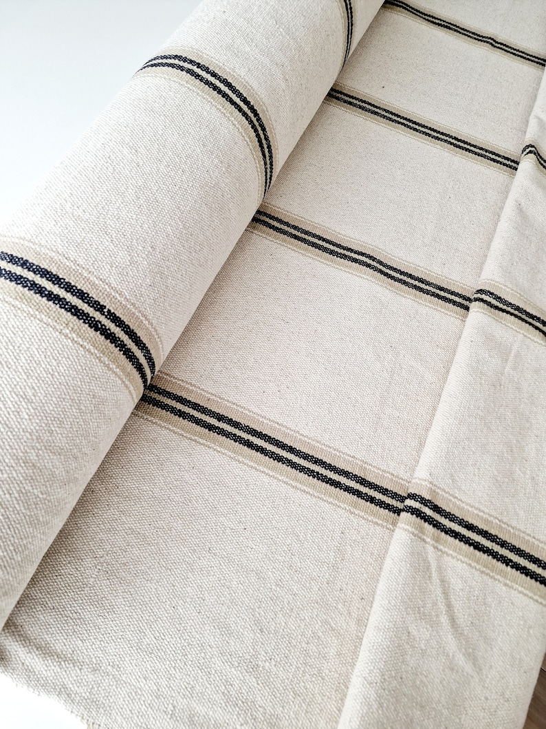 Pu&ograve; includere: Tessuto beige arrotolato con strisce orizzontali nere e beige. Il tessuto ha un aspetto strutturato, forse lino. Le strisce sono uniformemente distanziate sulla larghezza del tessuto, creando un design semplice e classico.