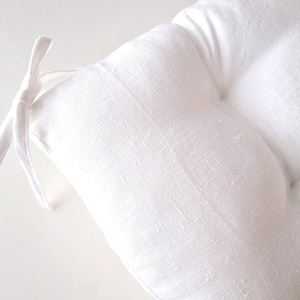 Close-up of a bright white linen chair cushion on a white background, highlighting the natural woven fabric texture. The image shows a corner of the plump, tufted seat pad with an integrated matching white linen tie for securing to a kitchen or dining chair.