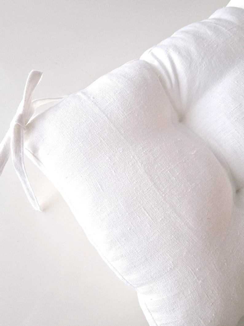 Close-up of a bright white linen chair cushion on a white background, highlighting the natural woven fabric texture. The image shows a corner of the plump, tufted seat pad with an integrated matching white linen tie for securing to a kitchen or dining chair.