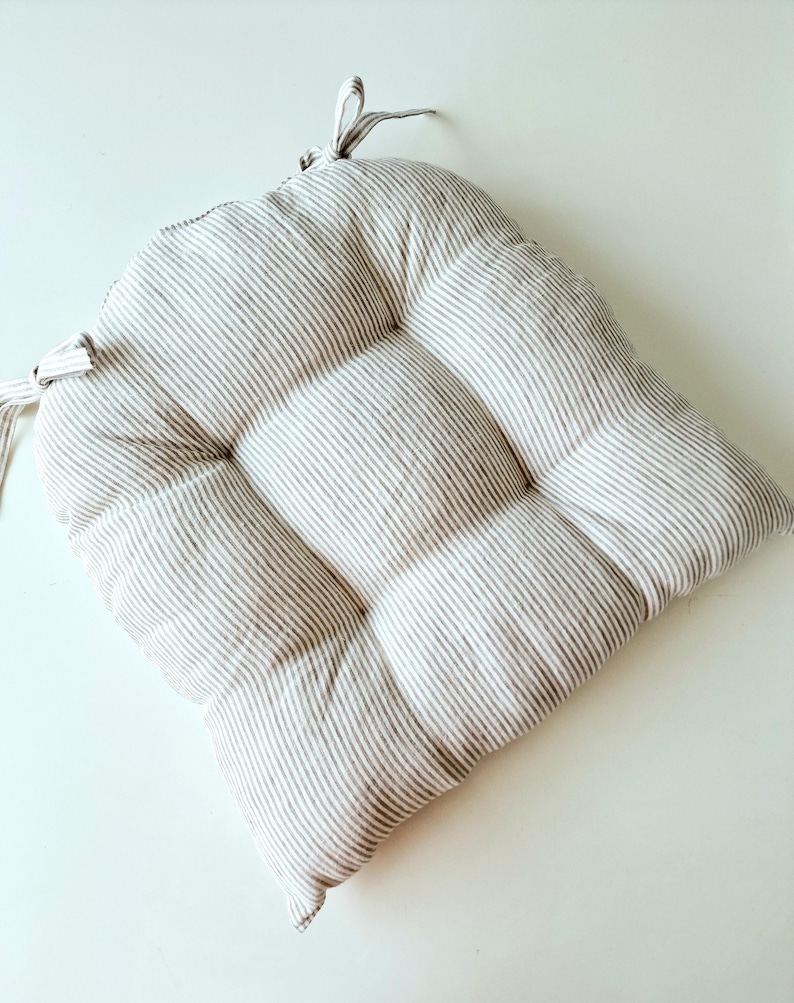 A square, tufted linen seat cushion with a grey and off-white striped pattern, placed flat on a clean white surface. The farmhouse style kitchen chair pad features a soft, plump filling and integrated fabric ties at the back corners for securing to a chair.