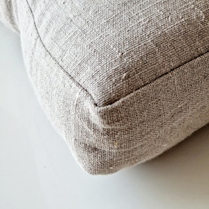 May include: Close-up of a square, light gray pillow with a textured, woven fabric. The pillow has a visible seam around the edges, and the fabric appears to be a natural fiber. The pillow is resting on a white surface.