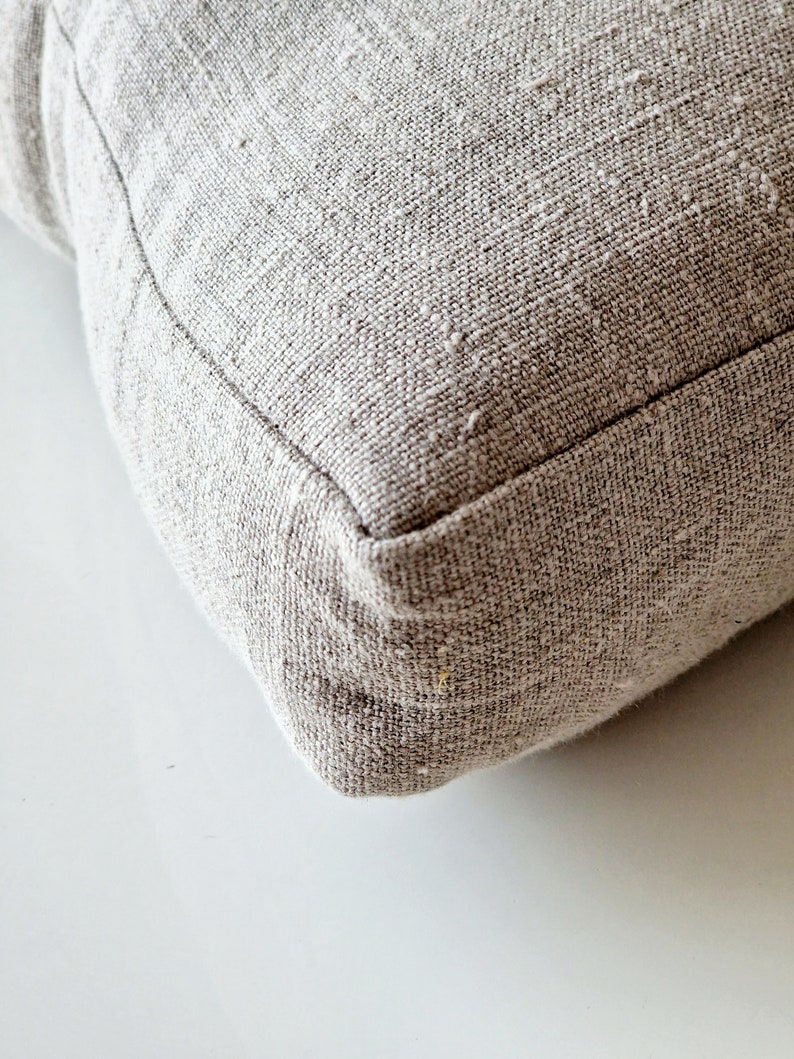 May include: Close-up of a square, light gray pillow with a textured, woven fabric. The pillow has a visible seam around the edges, and the fabric appears to be a natural fiber. The pillow is resting on a white surface.